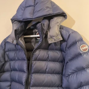 Blå Colmar pufferjacka med huva - Säljer en blå pufferjacka från Colmar med huva och dragkedja framtill. Jackan har två sidofickor med dragkedja och ett Colmar-märke på ärmen. Fodret är mörkgrått och jackan har en snygg, quiltad design som ger en cool look.