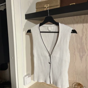 Vit ribbad väst med knappar - Vit ribbad väst från H&M med v-ringning och två knappar framtill. Västen har svarta kontrastsömmar längs kanterna och är ärmlös med en figurnära passform. Perfekt för lager-på-lager och enkel att styla.