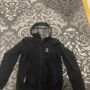 Haglöfs Jacka - Svart vindjacka från Haglöfs i Gore-Tex-material med huva och hel dragkedja. Jackan har två fickor med dragkedja och meshfoder på insidan. Perfekt för blåsiga och regniga dagar. Snygg och funktionell design med diskret logga på bröstet.