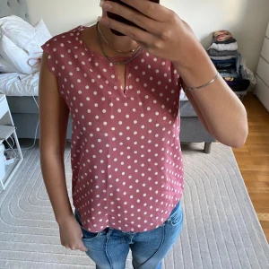 Rosa prickig blus med nyckelhålsringning - Söt rosa blus med vita prickar och en liten nyckelhålsringning framtill. Blusen är ärmlös med lätt volang vid axlarna och har en avslappnad passform. Perfekt att styla med jeans eller kjol för en trendig look.