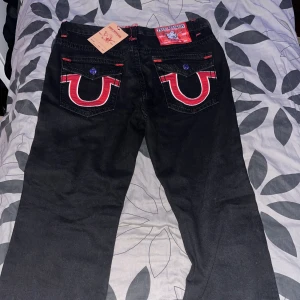 Svarta True Religion jeans med röd detalj - Svarta jeans från True Religion med ikoniska röda och vita broderade detaljer på bakfickorna. Jeansen har röda kontrastsömmar och klassisk femficksdesign. Märkespatch baktill och knappar med logga. Snygg och unik streetstil. Priset går å diskutera vid snabb affär 