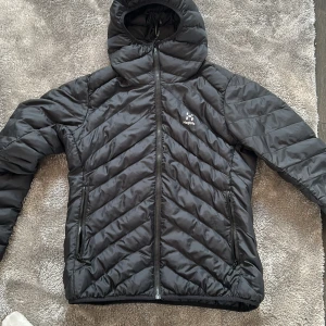 Svart pufferjacka från Haglöfs - Svart pufferjacka från Haglöfs med huva och snygga diagonala quiltade sömmar. Jackan har två dragkedjeförsedda fickor och hel dragkedja framtill. Perfekt för kalla dagar och har en clean look med diskret logga på bröstet.