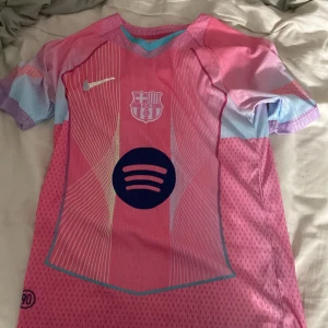FC Barcelona x Nike rosa fotbollströja S - Säljer en FC Barcelona fotbollströja från Nike i rosa och ljusblått med vita linjer och klubbmärke på bröstet. Tröjan har korta ärmar, Dri-FIT ADV-material och stor Spotify-logga framtill. Perfekt för dig som vill sticka ut på planen eller läktaren.