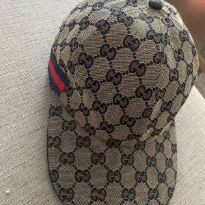 Gucci keps med GG-mönster och röd rand - Exklusiv keps från Gucci i beige med klassiskt GG-monogram över hela ytan. Kepsen har en böjd skärm, justerbar rem bak i svart skinn och en ikonisk röd och marinblå rand på sidan. Tillverkad i bomullscanvas med detaljer i skinn. Perfekt för dig som vill sticka ut. Kepsen är oanvänd och säljer på grund av brist på användning, skick 10/10