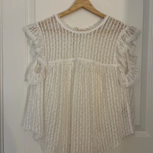 Vit spetsblus med volangärmar - Superfin vit blus i spets med vertikala ränder och volangdetaljer vid ärmarna. Blusen är ärmlös och har en luftig, flowy passform. Perfekt för dig som gillar romantisk och söt stil med mycket detaljer.