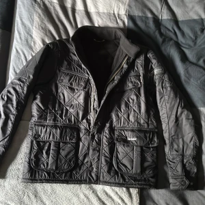 Svart quiltad jacka från Barbour - Svart quiltad jacka från Barbour International med snygga detaljer som tryckknappar, dragkedja och flera fickor. Jackan har hög krage med knäppning och klassiskt Barbour-märke på ärmen. Perfekt för dig som vill ha en stilren och funktionell look.
