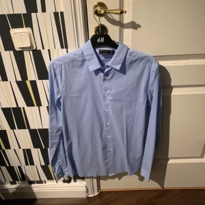 Ljusblå skjorta från ASOS DESIGN - Snygg ljusblå skjorta från ASOS DESIGN i klassisk modell med krage och knappar framtill. Skjortan har lång ärm och är tillverkad i mjuk bomull, perfekt för en clean och stilren look.