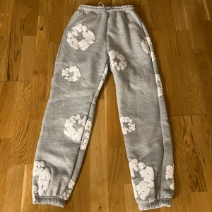 Grå mjukisbyxor Denim Tears med tryck - En grå mjukisbyxa med resår i midjan och vid bensluten. Byxan har ett mönster av vita molnformade tryck utspridda över hela tyget. Den ser bekväm och mysig ut, perfekt för  vardag,gym eller hemmabruk passar xxs och xs