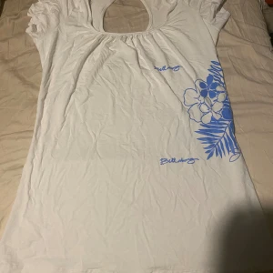 Vit blommig topp från Billabong - Vit topp från Billabong med blått blommönster och blad på ena sidan. Toppen har korta ärmar, rund halsringning med liten öppning i ryggen och är gjord i mjukt bomullsmaterial. Perfekt för en chill sommardag.