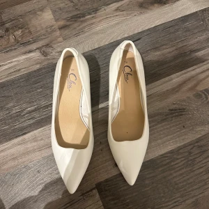 Vita pumps med spetsig tå från Clou - Snygga vita pumps från Clou i storlek 38. Skorna har en spetsig tå och är tillverkade i ett slätt syntetmaterial med låg klack. Perfekta för dig som vill ha en clean och stilren look till outfiten.