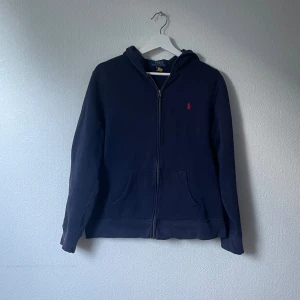 Mörkblå hoodie från Polo Ralph Lauren - Snygg mörkblå hoodie från Polo Ralph Lauren i storlek XL i barn. Tröjan har huva, dragkedja framtill och två praktiska fickor. Klassisk röd Polo-logga broderad på bröstet. Perfekt för chill dagar och enkel att matcha med jeans eller joggers.