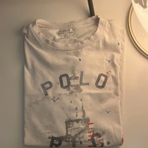 Vit t-shirt Polo Ralph Lauren L - Vit t-shirt från Polo Ralph Lauren i storlek L i barn så motsvarar xs men även kanske s. Tryck framtill med texten POLO R.L.C. och motiv av en fyr med fåglar. Klassisk rund halsringning och korta ärmar. Skön och lätt i bomull, perfekt för en chill stil. 