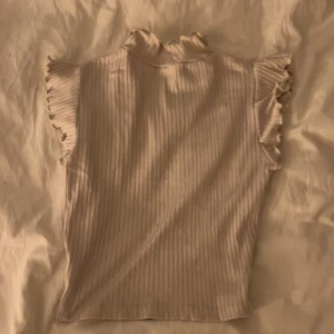 Beige ribbad topp med volangärm - Säljer en ljusbeige ribbad topp med volangdetaljer på ärmarna och liten ståkrage. Toppen är ärmlös med volang och har en mjuk, stretchig känsla. Perfekt att styla med jeans eller kjol för en trendig look. Knappt använd
