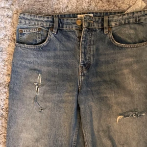 Blå slitna raka jeans - Säljer ett par blå jeans från Zara med raka ben och flera slitningar på både lår och knän. Jeansen har klassisk femficksdesign, normal passform och är tillverkade i bomull. Perfekta för en avslappnad och trendig look.