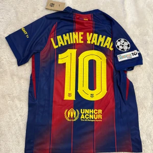 FC Barcelona Lamine Yamal #10 matchtröja - Säljer en officiell FC Barcelona fotbollströja med Lamine Yamal och nummer 10 på ryggen. Tröjan är randig i mörkblått och rött med gula detaljer, Champions League-märke på ärmen och sponsorer som Spotify och UNHCR. Materialet är lätt och andas, perfekt för match eller träning.