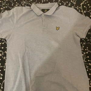 Ljusblå pikétröja från Lyle & Scott - Snygg ljusblå pikétröja från Lyle & Scott med klassisk krage och korta ärmar. Tröjan har gul broderad logga på bröstet och knappar vid halsen. Perfekt för dig som gillar stilrena och enkla plagg med en sportig touch.