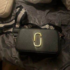 Svart axelväska Marc Jacobs - Säljer en svart axelväska från Marc Jacobs med guldiga detaljer och bred axelrem med logga. Skriv för mer information om äkthet! Väskan har två dragkedjor och är tillverkad i ett tåligt skinnliknande material. Perfekt storlek för det viktigaste och riktigt snygg till streetwear eller casual outfits.