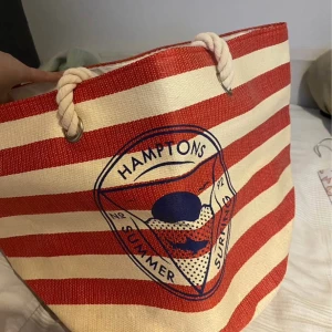 Strandväska - Stor och rymlig strandväska med röda och vita ränder, grova rep-handtag och tryckt Hamptons-motiv framtill. Perfekt för stranden eller sommarens äventyr. Tillverkad i slitstark textil med marint inspirerad design. Köpt från stadium med prislapp.