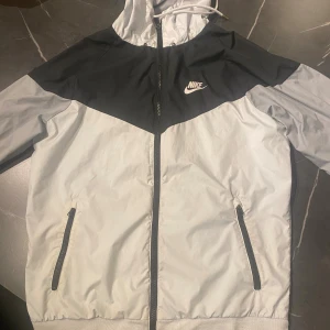 Nike svartvit vindjacka med huva S - Nike vindjacka i svart och vitt med dragkedja framtill, två fickor med dragkedja och justerbar huva. Jackan har klassisk Nike-logga på bröstet och är tillverkad i lätt polyester. Perfekt för dig som gillar sportig och clean stil.