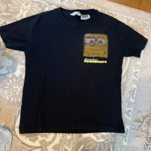 Svart SpongeBob SquarePants t-shirt - Svart t-shirt med färgglatt SpongeBob-motiv på bröstet och stor vit SpongeBob-tryck på ryggen. Klassisk passform och rund hals. Tillverkad i mjuk bomull, perfekt för dig som älskar tecknade serier och vill sticka ut med en ikonisk look.vet ej storlek .det kan vara mellan 7-8 år märken på bröstet är liten riven på själva kant ,som man ser på bild fyra,