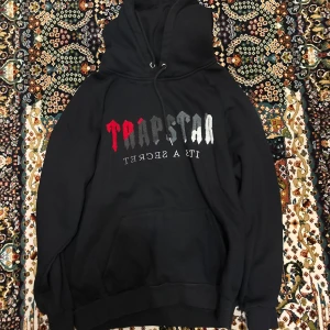 Svart Trapstar hoodie med tryck - Svart hoodie från Trapstar med stor logga i rött, grått och vitt framtill. Klassisk känguruficka, snörning i huvan och mjukt material. Perfekt för dig som gillar streetwear och vill sticka ut med en fet logga.