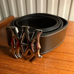 Amiri bälte - Snyggt svart Amiri bälte i äkta skinn med grov textur och vita sömmar längs kanterna. Spännet är stort och i blankt silver med sin ikoniska logotyp i en cool, edgy design. Perfekt för att lyfta din outfit med en streetweare accessoar. Helt nytt!