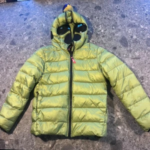 Grön skidjacka AI Riders 12 år - Grön skidjacka från AI Riders on the Storm med hel dragkedja och unik huva med inbyggda svarta goggles (något repiga) Jackan har en gul och blå tofs på huvan och är tillverkad i glansig polyester. Storlek 12 år.