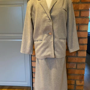 Beige Dräkt med Kavaj och Kjol. Strl 40 från 1980-talet - Klassisk dräkt i en tidlös design. Kavajen har en enkelknäppning, diskreta fickor och en smickrande passform. Kavajen är Helfordrad. Kjolen är en rak modell med en elegant slits baktill och ett matchande bälte. 20% Ull 5% Nylon 35% Akryl  30% Polyester  10% O.F Tillverkad på 1980-talet Från Italien Skärpet i kjolen medföljer