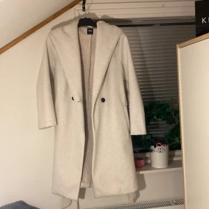 Vit kappa med luva från Zara - Stilren beige kappa från Zara med luva och bälte i midjan. Kappan har lång ärm, knäppning framtill och en mjuk, lite ullig känsla. Perfekt för dig som vill ha en clean look med snygg passform och extra cozy känsla.