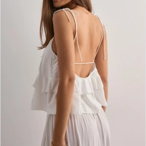 Vit volangtop med knytband - Supersöt vit linnetopp med dubbla volanger och tunna axelband som knyts på axlarna. Toppen är croppad och har öppen rygg med smala band som går över ryggen. Perfekt för varma dagar och ger en luftig känsla.