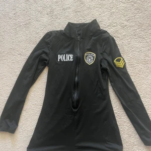 Svart polis klänning med dragkedja - Helt ny aldrig använd Polisdräkt till haloween med lång ärm och dragkedja framtill, dekorerad med broderade polispatchar och POLICE-tryck. Medföljer svarta bälten för extra detalj. Perfekt för temakväll eller maskerad! Materialet känns som polyester och har en slim passform.