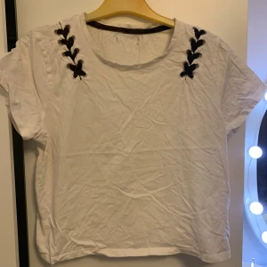 Vit t-shirt med snörning vid axlarna - Säljer en vit t-shirt med korta ärmar och cool snörningsdetalj i svart vid båda axlarna. Rund halsringning och loose passform, perfekt för en chill och avslappnad stil. Materialet känns som bomull och toppen är enkel att matcha med jeans eller shorts.