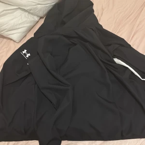 Svart vindjacka från Under Armour YXL - Svart vindjacka från Under Armour med vit logga på bröstet och vita detaljer längs ärmarna. Jackan har huva, hel dragkedja och meshfoder på insidan för extra ventilation. Perfekt för sport och träning när du vill ha en clean och sportig look. Aldrig använd!