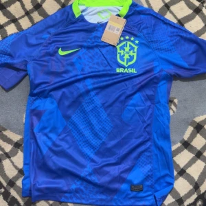 Brasilien fotbollströja - Snygg blå Brasilien fotbollströja från Nike med neongula detaljer och tryckt klubbmärke på bröstet. Tröjan har korta ärmar och ett mönster i olika blå nyanser. Perfekt för dig som vill sticka ut på planen eller heja på landslaget.