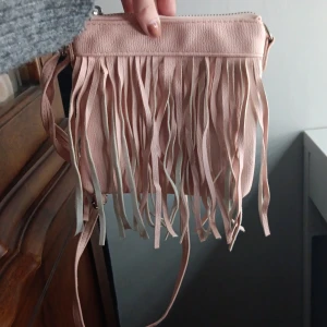 Rosa axelväska med fransar - Säljer en ljusrosa axelväska i syntetmaterial med långa fransar framtill. Väskan har dragkedja upptill och en smal axelrem. Perfekt för dig som vill ha en cool och trendig accessoar med lite boho-vibe.
