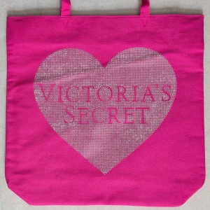 Rosa shoppingväska Victoria's Secret - HELT OANVÄND SHOPPINGVÄSKA. Stor och rymlig rosa shoppingväska från Victoria's Secret med ett glittrigt hjärta och logga på framsidan. Väskan har två handtag och svart innerfoder. Perfekt för att bära med sig allt man behöver under dagen.