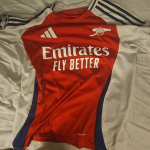 Arsenal Saka 7 kortärmad matchtröja - Riktigt snygg Arsenal fotbollströja med Saka 7 på ryggen. Tröjan är kortärmad med röd framsida, vita ärmar och blå detaljer. Adidas-logga, klubbmärke och Champions League-patch på ärmen. Materialet är lätt och andas, perfekt för match eller träning.