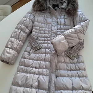 Ljusgrå lång dunjacka med fuskpäls - Snygg ljusgrå lång dunjacka från Ableboy med quiltad design och avtagbar huva med fuskpäls. Jackan har två snedställda fickor med spännen och dragkedja framtill. Perfekt för kalla dagar och ger en trendig vinterlook.