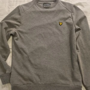 Grå sweatshirt från Lyle & Scott - Snygg grå sweatshirt från Lyle & Scott med klassisk rund hals och gul logga på bröstet. Tröjan har ribbade muddar vid ärmslut och nederkant..