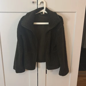 Mörkgrön teddyjacka i storlek S - Mysig mörkgrön teddyjacka med dragkedja framtill och två fickor. Jackan har hög krage och är tillverkad i ett fluffigt teddy-material som håller dig varm under kyliga dagar. Perfekt för dig som gillar en chill och trendig look.