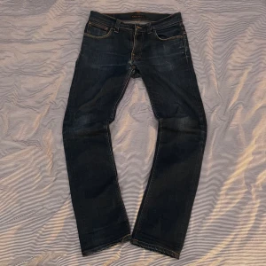 Nudie Jeans - Snygga mörkblå Nudie jeans av modellen Tape Ted. Dom har snygga slitningar som ger en tidlös look. Jeansen har en liten slitning i skrevet, men syns knappt när man har på sig dom. Jeansen går även att lämna in på lagning helt gratis i vilken Nudie-butik som helst ifall de skulle gå sönder. Hör av er om ni har några funderingar!