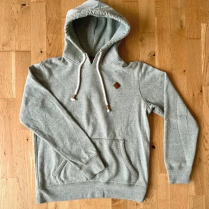 Grå hoodie från Jack & Jones - Säljer en stilren grå hoodie från Jack & Jones med huva och vita snören. Tröjan har en klassisk känguruficka framtill och en liten brun logodetalj på bröstet. Tillverkad i mjuk bomulls- och polyestermix för extra komfort.