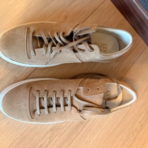 Taupe mockasneakers från Jim Rickey - Snygga taupefärgade sneakers från Jim Rickey i modellen Cloud. Skorna är gjorda i mjuk mocka med vita gummisulor och har klassisk snörning. Perfekta för dig som gillar stilrena och minimalistiska sneakers med en modern touch. Endast provade, nyskick!