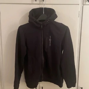 Svart fleecejacka från Sail Racing - Svart fleecejacka från Sail Racing med huva och dragkedja framtill. Jackan har en bröstficka med dragkedja och logga på både bröstet och huvan. Mjuk insida och långärmad modell, perfekt för kyliga dagar.
