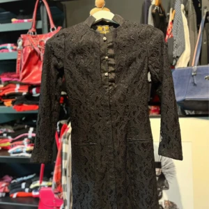 Svart spetsklänning Miss Perada - Svart klänning från Miss Perada med elegant spetsmönster över hela klänningen. Den har långärmade ärmar, ståkrage och dekorativa knappar framtill. Klänningen går att öppna hela vägen pga knappar framtill som ej är synliga.