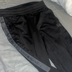 Svarta EA7 Emporio Armani mjukisbyxor - Svarta mjukisbyxor från EA7 Emporio Armani med elastisk midja och logga längs sidorna. Byxorna har en sportig look med smal passform och är tillverkade i ett mjukt, syntetiskt material som känns skönt mot huden. Perfekta för en chill och stilren outfit.