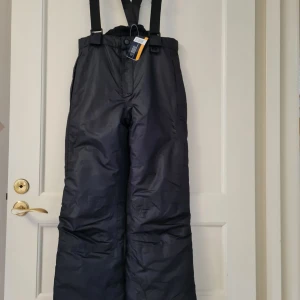 Svarta täckbyxor från Crivit Sports strl 146/152 10-12 år - Svarta täckbyxor från Crivit Sports med justerbara hängslen och dragkedja vid benslut. Byxorna har elastisk midja och är tillverkade i slitstarkt syntetmaterial, perfekta för kalla dagar. Passar dig som vill ha funktionella och bekväma vinterbyxor.