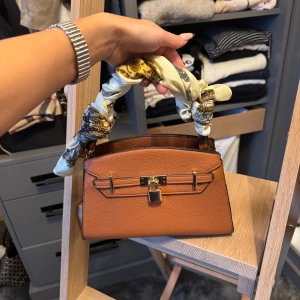 Brun-orange handväska med scarf-detalj - Snygg brun handväska i skinn med guldfärgade metalldetaljer och lås framtill. Handtaget är dekorerat med en mönstrad scarf i beige och gult. Väskan har dragkedja baktill och flera fack inuti för smart förvaring. Perfekt accessoar för att lyfta din outfit. Knapptanvänd och köpt i Santotini för 2 år sedan