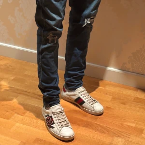 Gucci Ace sneakers stl 44 - Gucci Ace sneakers i vitt skinn med ikoniska gröna och röda ränder på sidan samt broderad orm i rött och svart. Platt sula med unikt mönster undertill, snörning och rund tå. Grön och röd detalj på hälen. Perfekt för dig som vill sticka ut med lyxig streetstyle. Fråga på!
