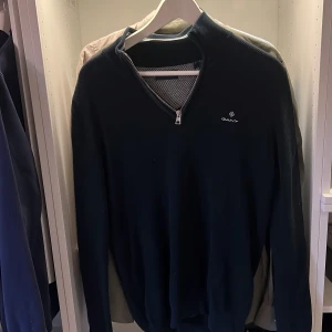 Mörkblå stickad tröja från Gant - Snygg mörkblå stickad tröja från Gant med halv dragkedja och broderad logga på bröstet. Tröjan har lång ärm och ribbade muddar, perfekt för lager-på-lager under höst och vinter. Klassisk och stilren design som funkar till det mesta.